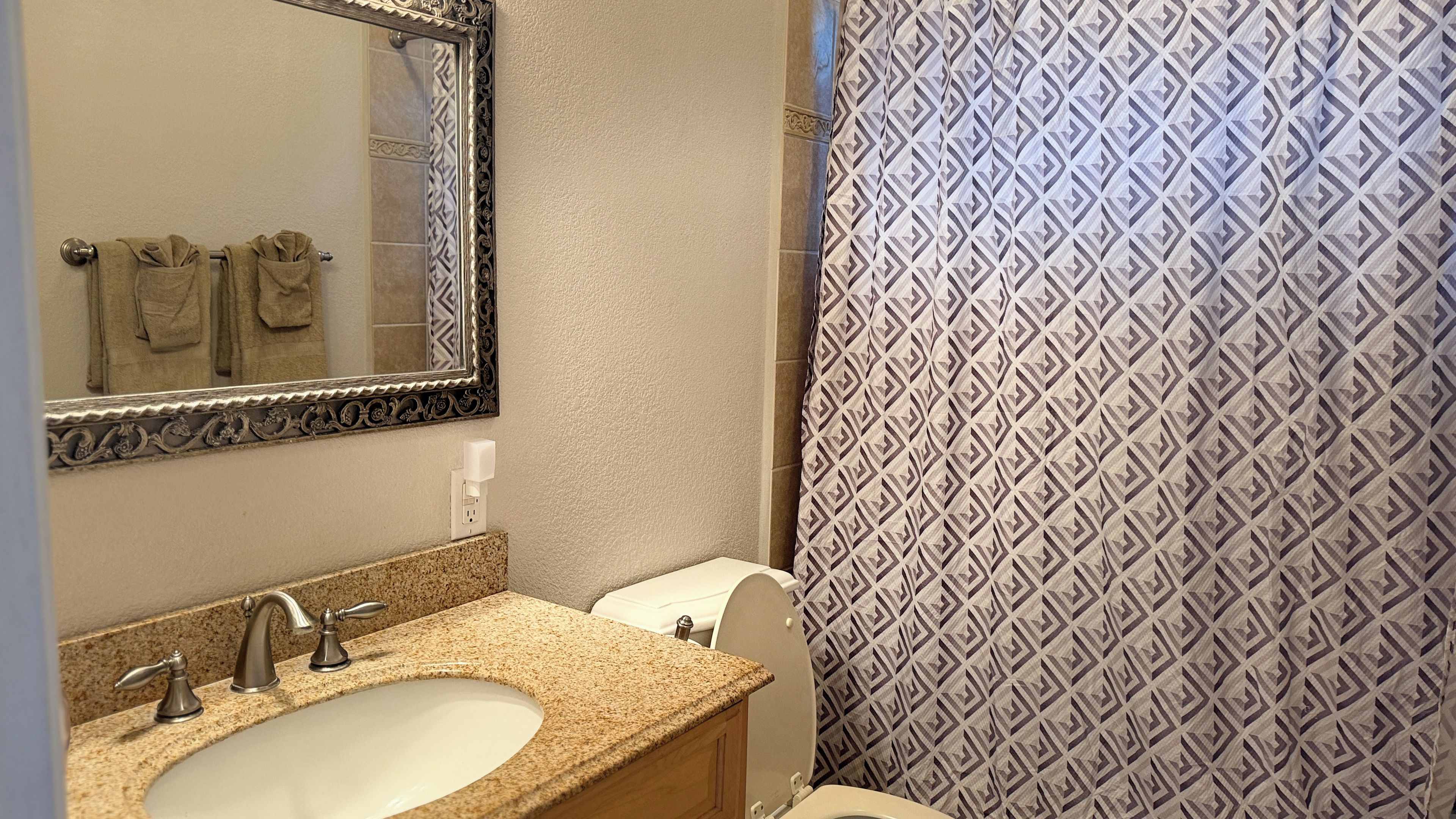 Un baño con lavamanos de encimera de granito, un espejo con marco decorativo, inodoro y cortina de ducha estampada.