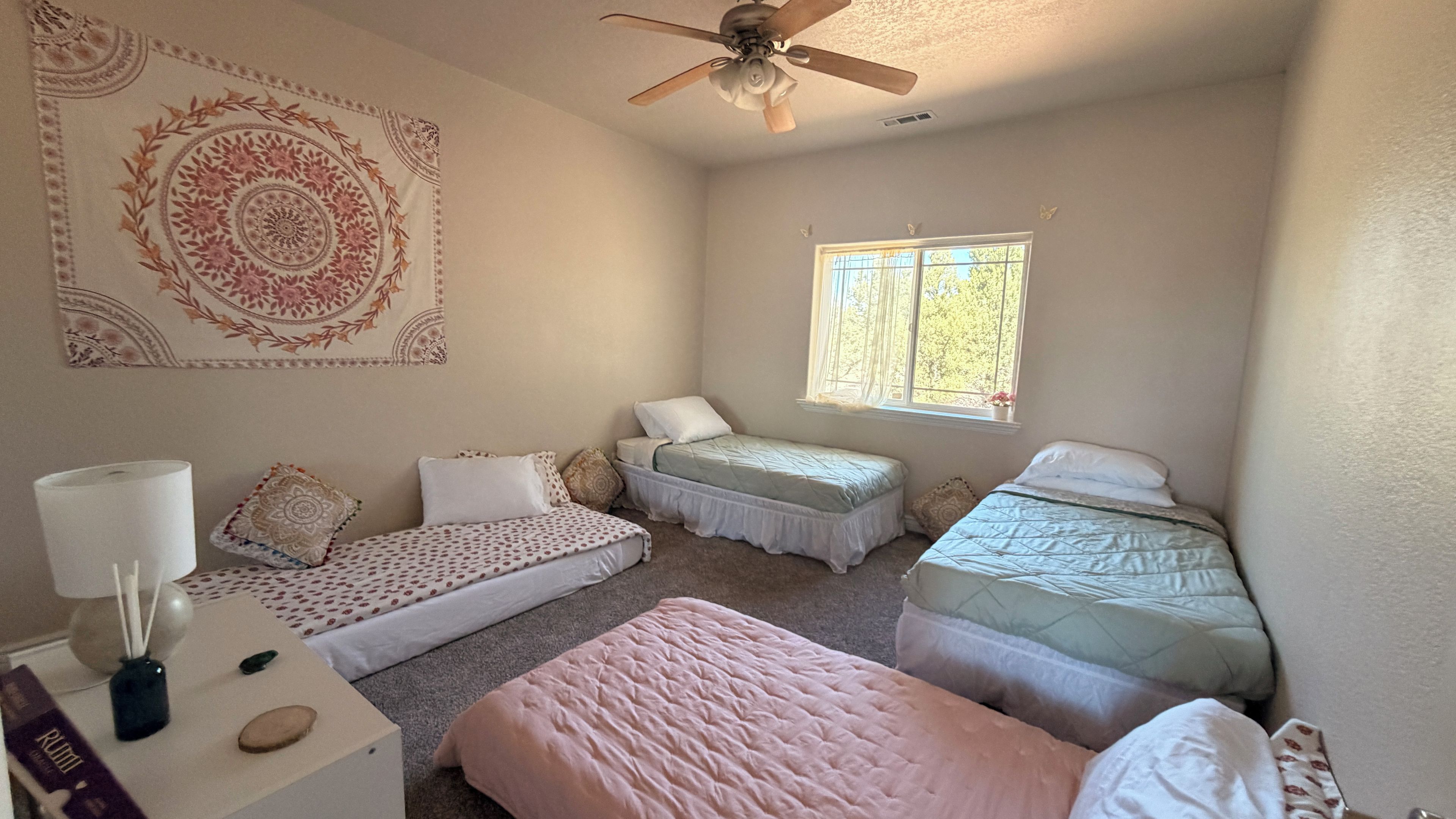 Quarto com duas camas de solteiro, ventilador de teto, janela com cortinas e tapeçaria na parede.