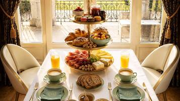 Une table de petit-déjeuner avec un présentoir à étages garni de viennoiseries, de fruits et de fromages, une sélection de café et de jus, et une vue sur un jardin par la fenêtre.