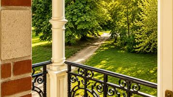 Un balcon doté d’une balustrade en fer forgé, d’un mur en briques et offrant une vue sur un parc verdoyant, avec des arbres et un sentier.