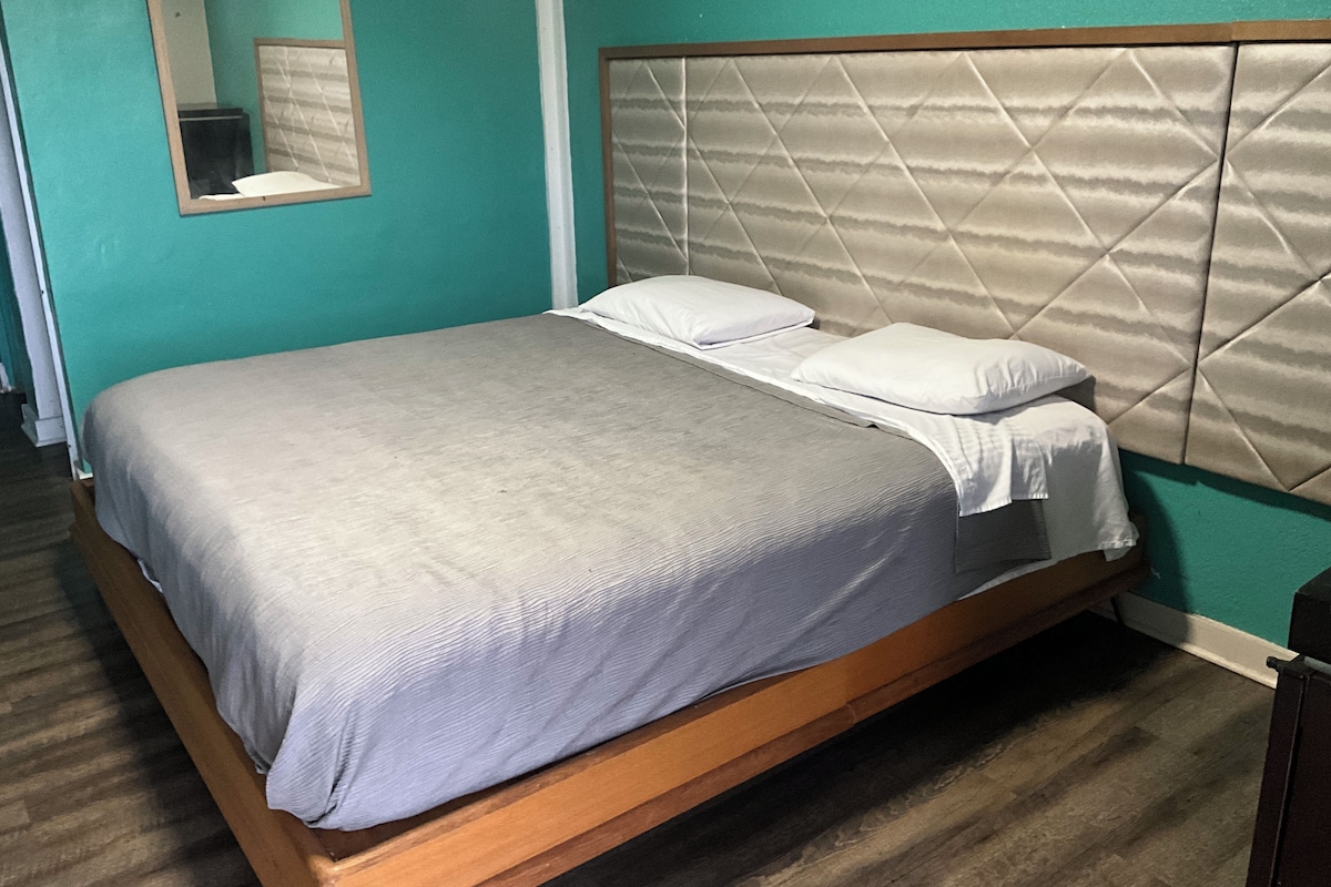 Uma cama de solteiro com estrutura de madeira e cabeceira acolchoada, posicionada em frente a paredes cor de verde-água e um espelho acima.