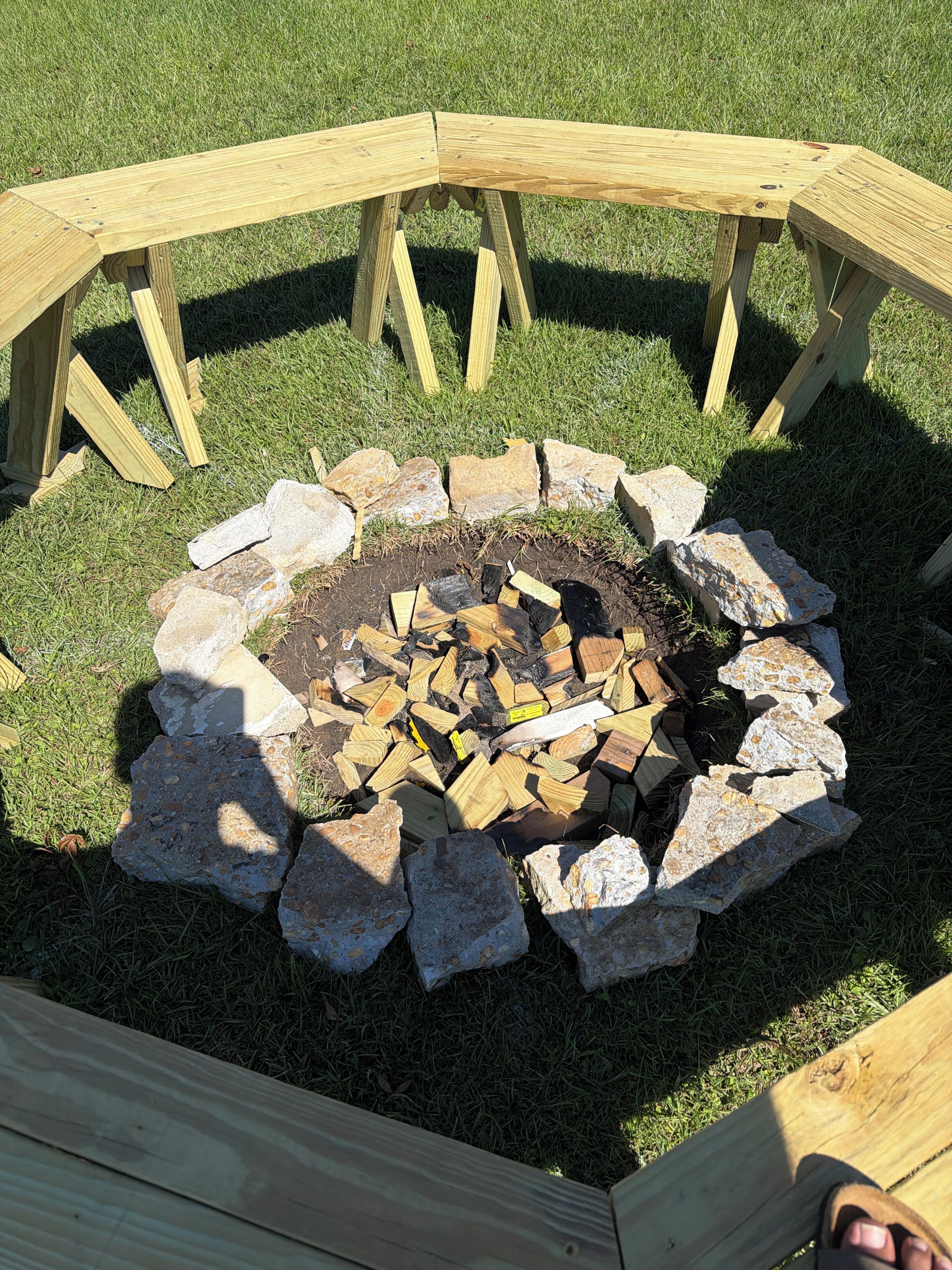 A circular fire pit with a wooden frame, surrounded by rocks and logs, set on a grassy lawn.