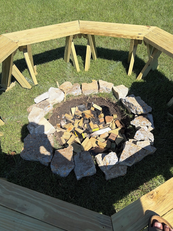 A circular fire pit with a wooden frame, surrounded by rocks and logs, set on a grassy lawn.