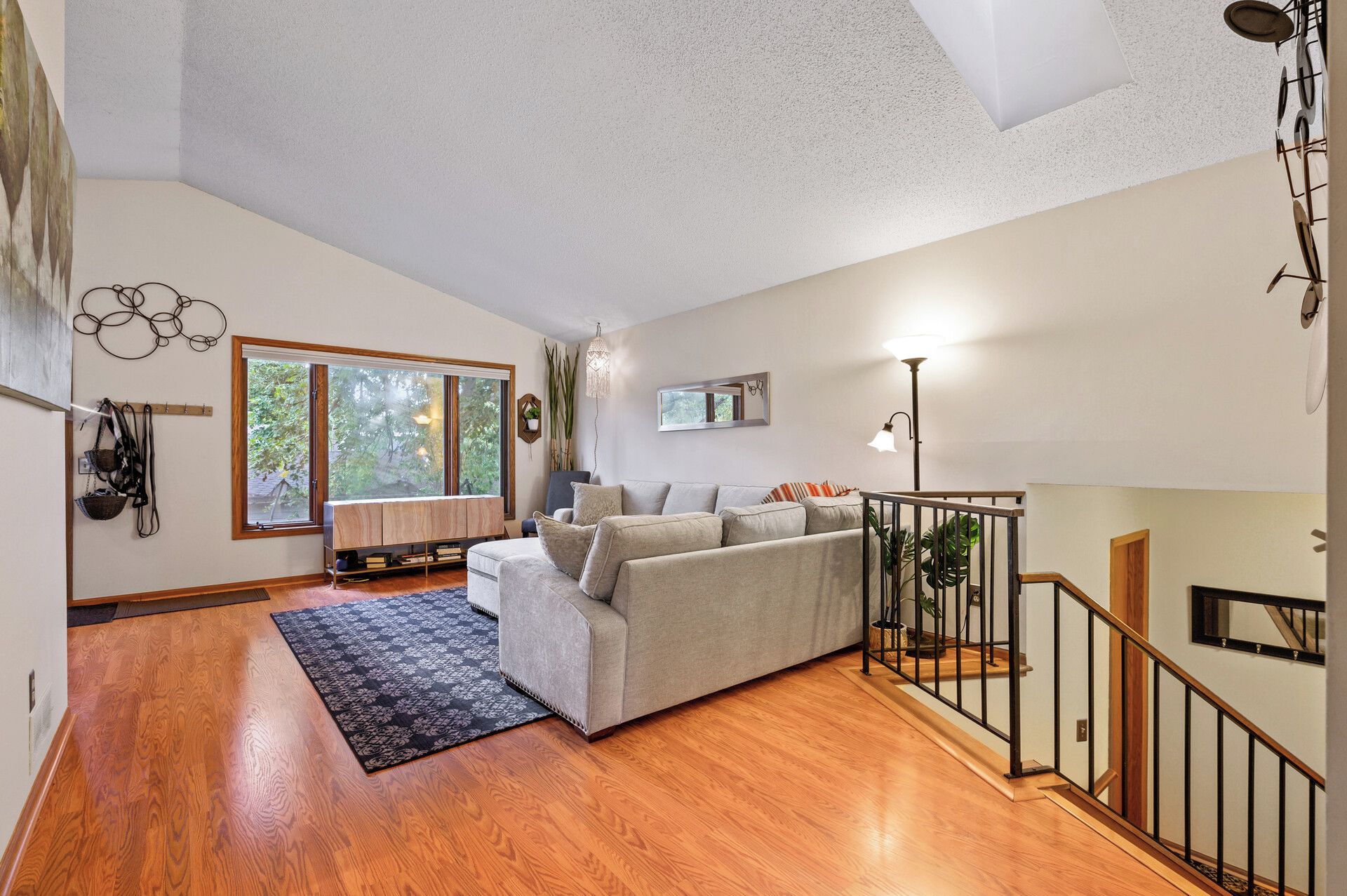 A spacious living area with a wooden floor, a grey sofa, a large window, and a staircase.