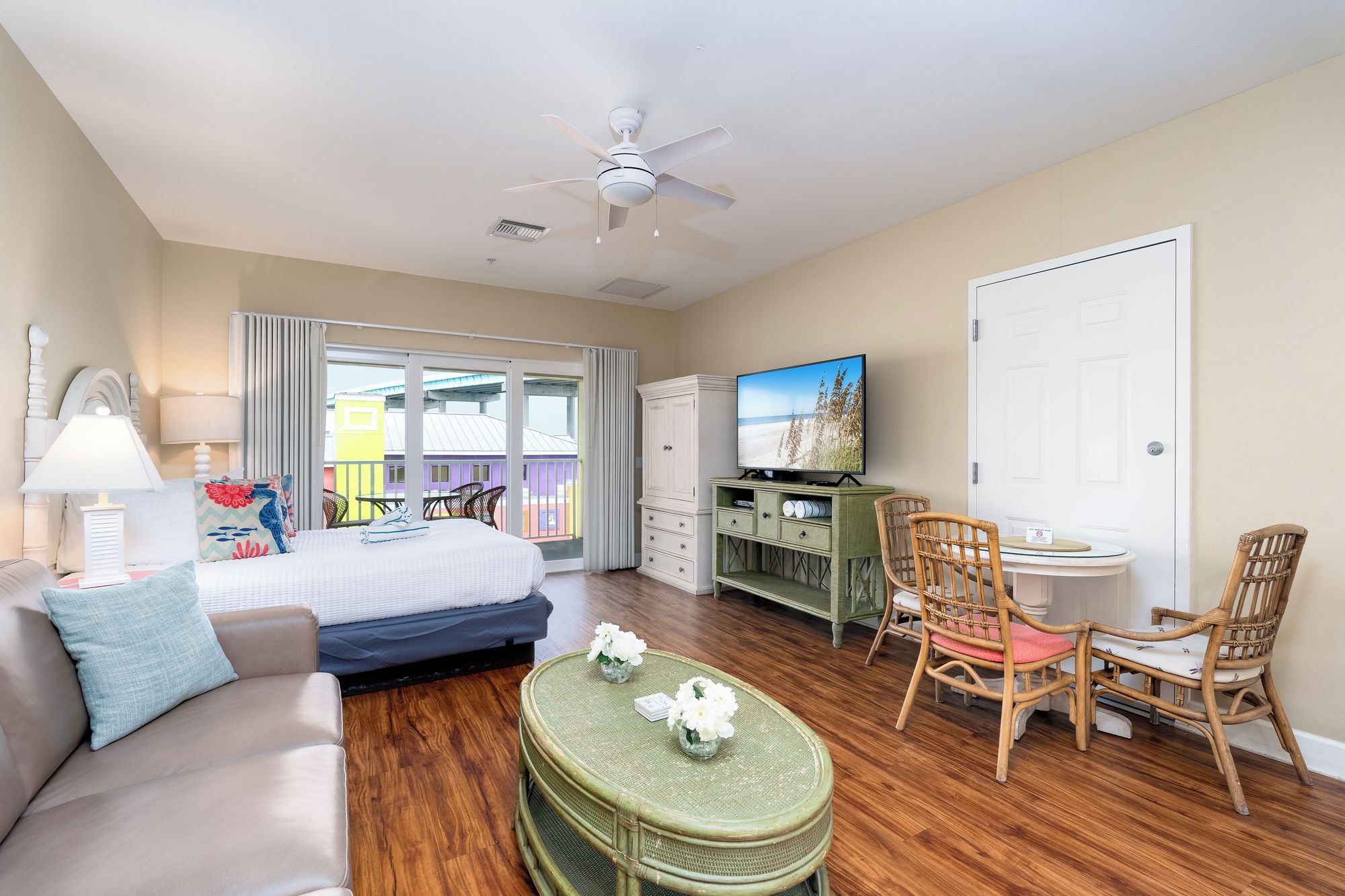 A hotel room with a bed, a sofa, a television, a dining table with chairs, and a balcony with a view of buildings.