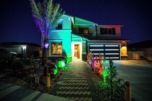 Une maison moderne à deux étages, dotée d’un garage, illuminée de lumières colorées et entourée de jardins aménagés.