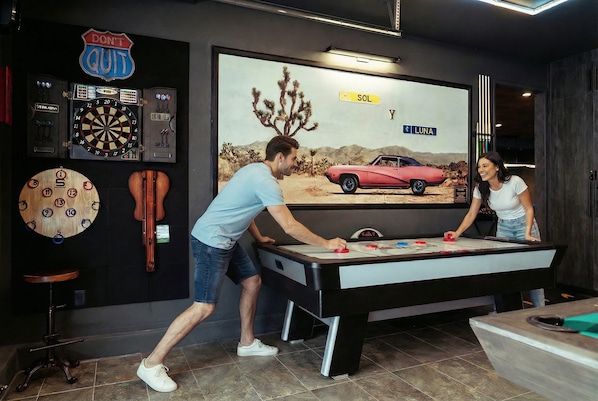 Une table de billard dans une pièce où l’on trouve également un jeu de fléchettes, du matériel de billard et une affiche représentant une voiture rouge.