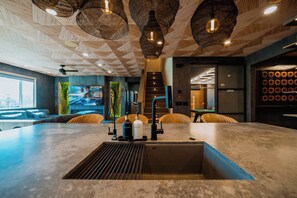 Un espace bar moderne doté d’un grand comptoir, d’un évier et de luminaires suspendus en osier.