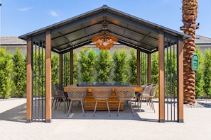Un espace extérieur aménagé, abrité et doté d’un barbecue, entouré de verdure et agrémenté d’un palmier.