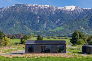 A modern house with a glass facade and a small porch, set against a backdrop of snow-capped mountains and a green valley. - Bay Paddock - Kaikoura Holiday Home (Kaikoura)