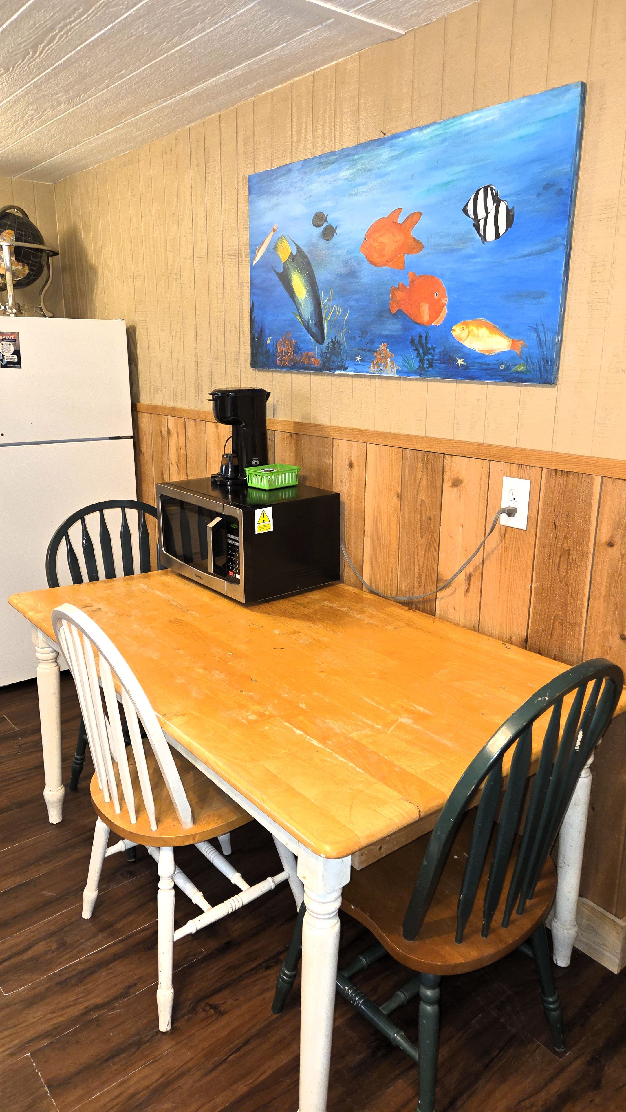 A dining area with a wooden table and chairs, a painting of fish on the wall, and a black appliance on the table.