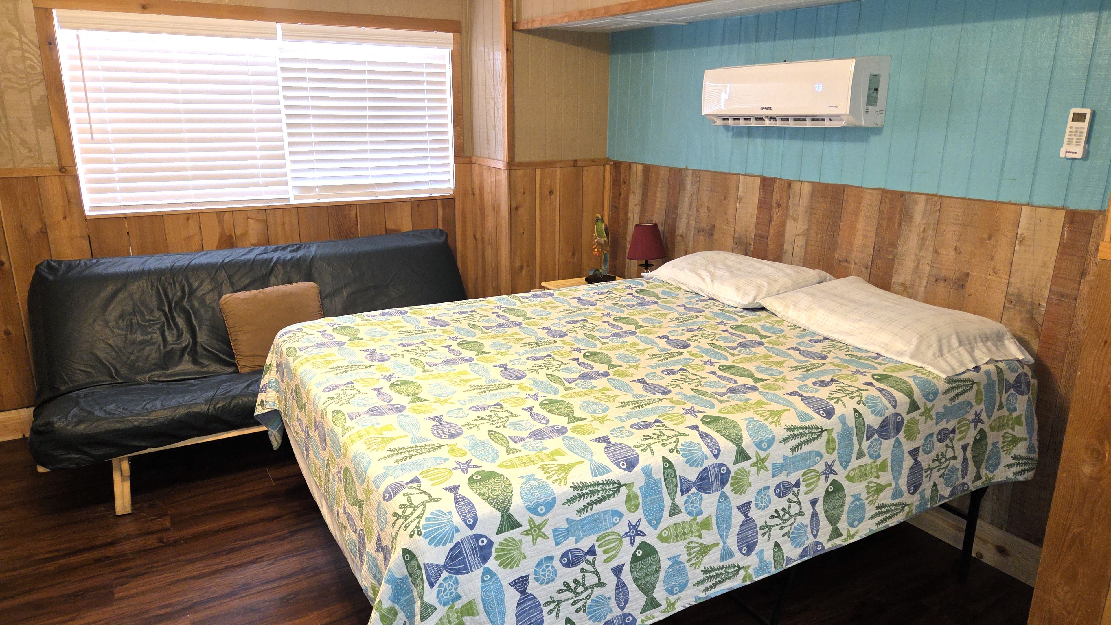 A room with a bed, a sofa, an air conditioning unit, and a window with blinds.