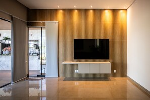 A modern interior with a large flat-screen TV mounted on a wall, a glass door leading to a balcony, and a shiny tiled floor. - Ch Cara com Piscina Hidro e Lago em Trindade - GO (Goiânia)