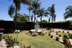 A landscaped garden with a central fountain, palm trees, and a hedge. - Ch Cara com Piscina Hidro e Lago em Trindade - GO (Goiânia)