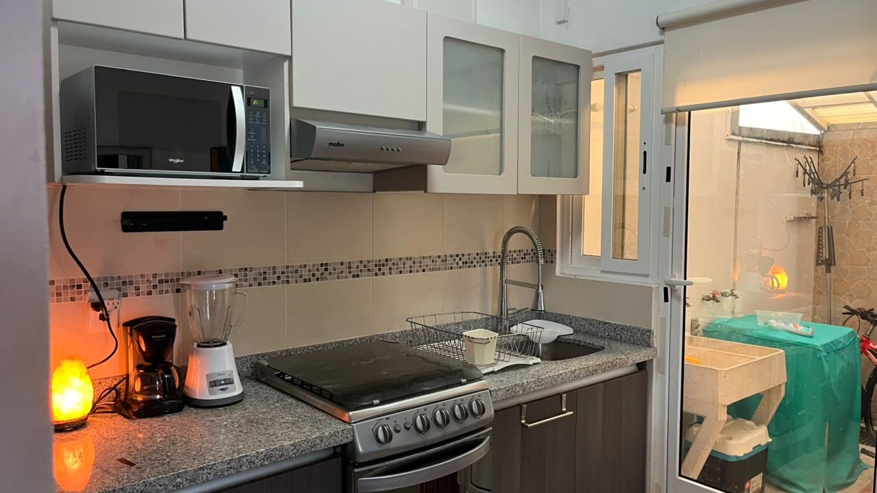 A compact kitchen with a microwave, stove, and blender on the countertop, a small sink, and a cabinet with a glass door.