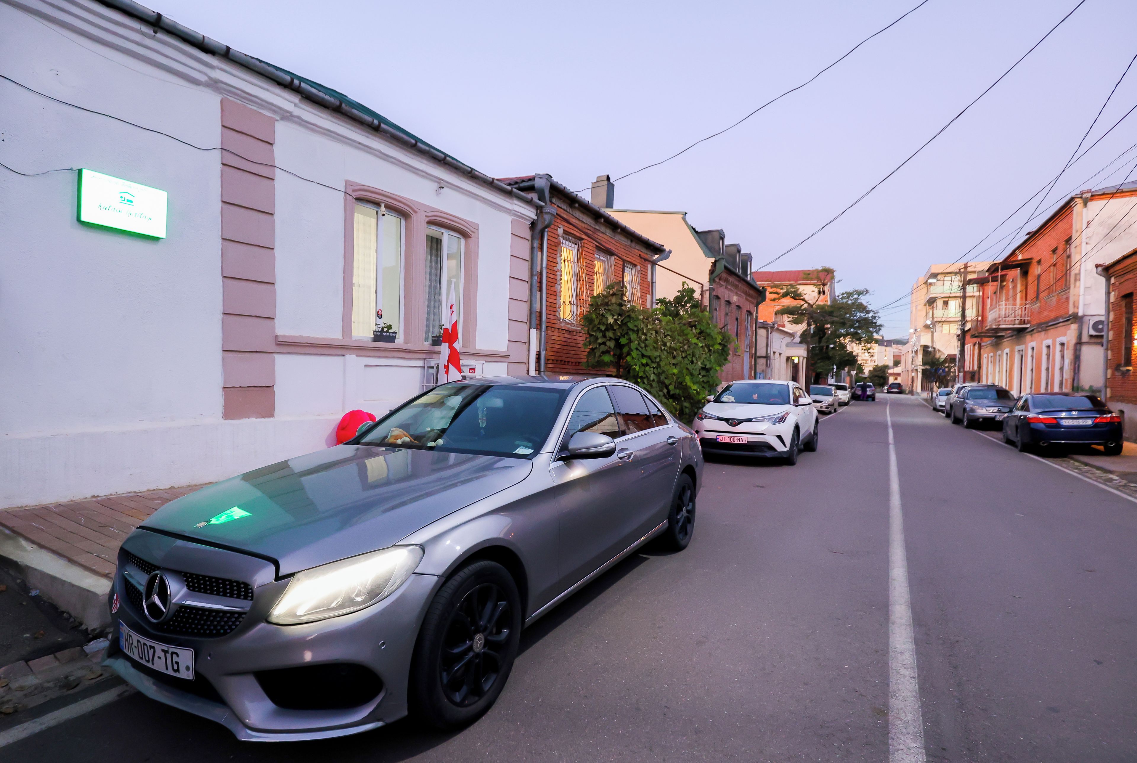 Rua com carros estacionados, incluindo um Mercedes-Benz prateado.