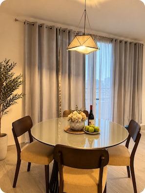 A dining area with a round glass table, two chairs, a potted plant, a vase with flowers, a bottle of wine, and a plate with apples. - Casco View Apartments & Rooftop Pool (Panamá)