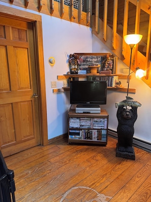Una puerta de madera, un televisor sobre un mueble bajo, una estantería con DVD y una estatua de oso con un pez en la cabeza.