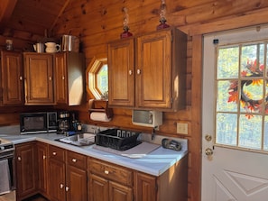A compact kitchen with wooden cabinets, a microwave, a coffee maker, and a small window with a view of trees. - LOG CABIN • 2-bedroom + loft. Access to private association waterfall + courts (Thornton)