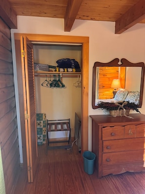 Un armario de madera con estantes, una barra para colgar ropa y un banco. A la derecha, un aparador con espejo y una lámpara.