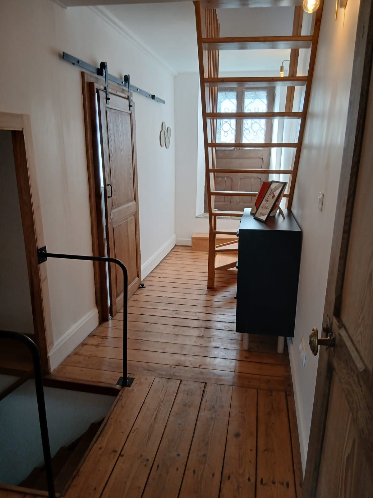 Uma escada de madeira com corrimão, uma porta com sistema deslizante e uma pequena mesa com moldura de quadro.