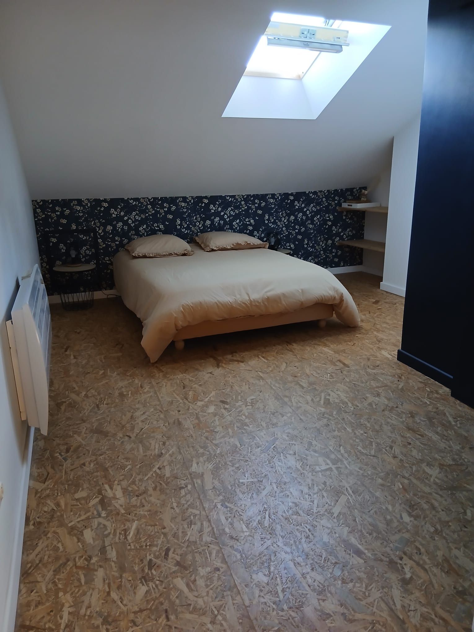 A bedroom with a sloped ceiling, a skylight, a bed with a beige cover, and a wall with a floral pattern.