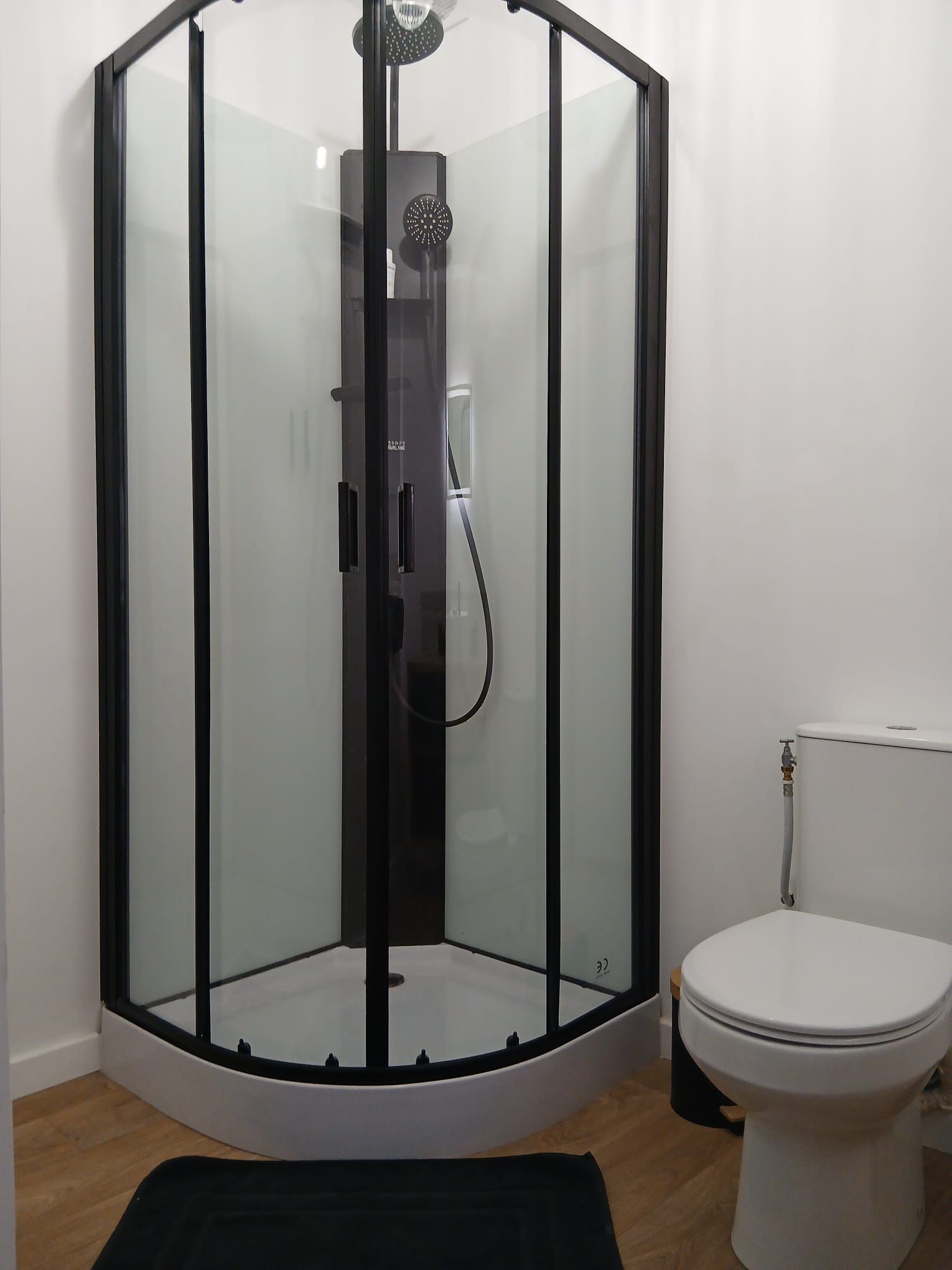 Een douche met glazen wanden en een zwart frame, een wit toilet en een houten vloer.
