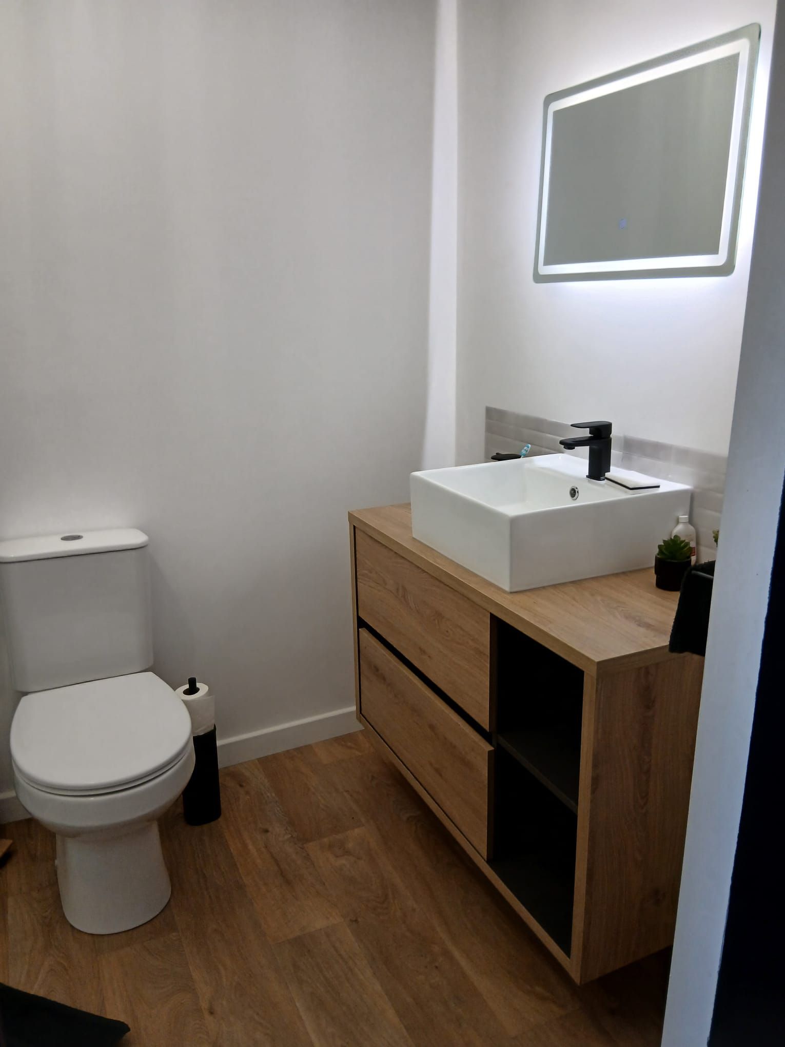 Ein modernes Badezimmer mit Toilette, einem weißen Waschbecken mit schwarzem Wasserhahn und einem hölzernen Waschtisch mit Schubladen.