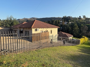 A residential property with a fenced yard, a house with a tiled roof, and a hilly landscape in the background. - Mountain Drive Apartments (Mbabane)