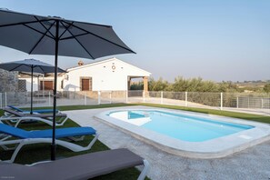 A swimming pool with lounge chairs and umbrellas, surrounded by a white fence and a building with a tiled roof. - Vrbo Property (Coin)