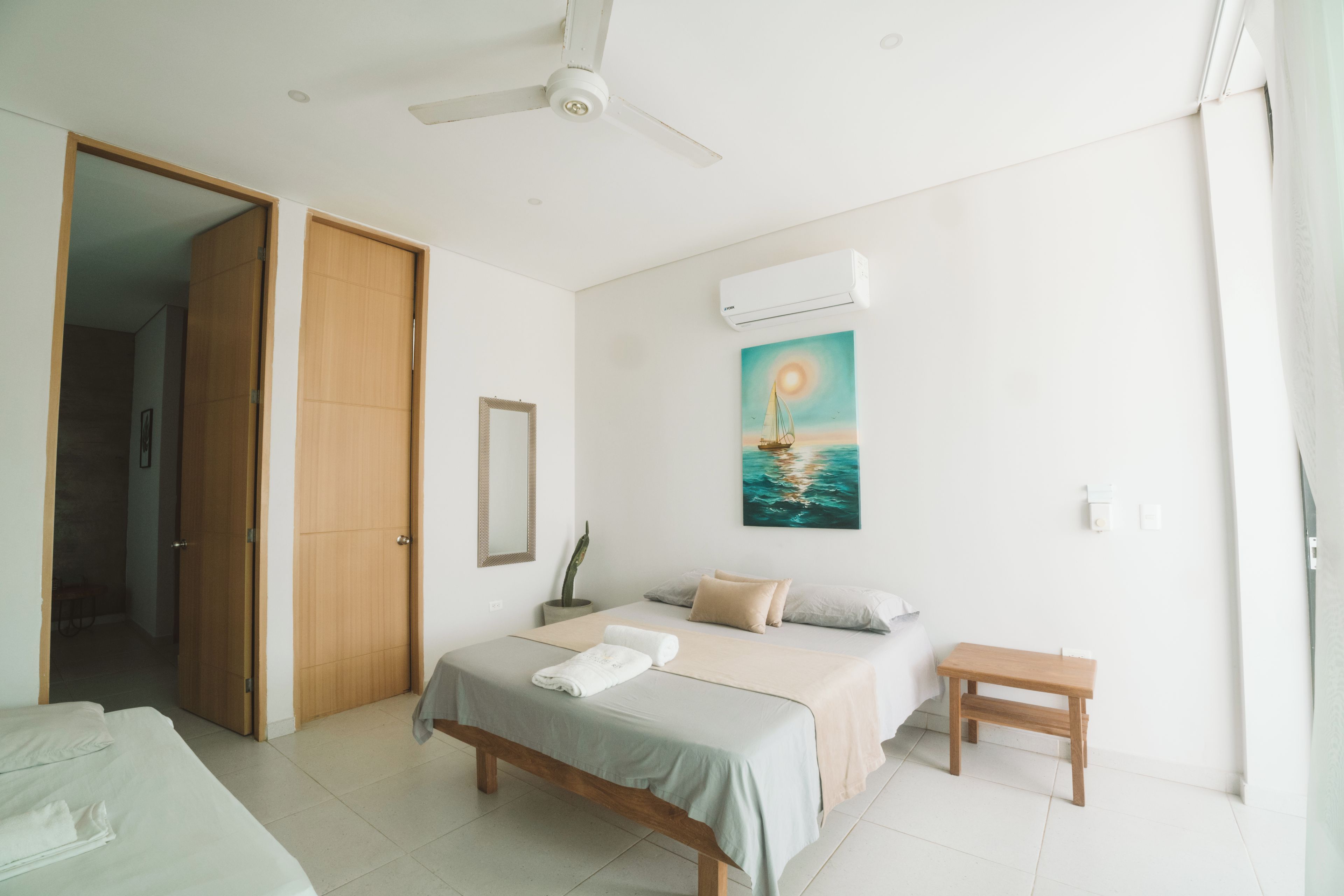 A bedroom with a bed, a bedside table, a ceiling fan, an air conditioning unit, a painting of a sailboat, and a door leading to another room.