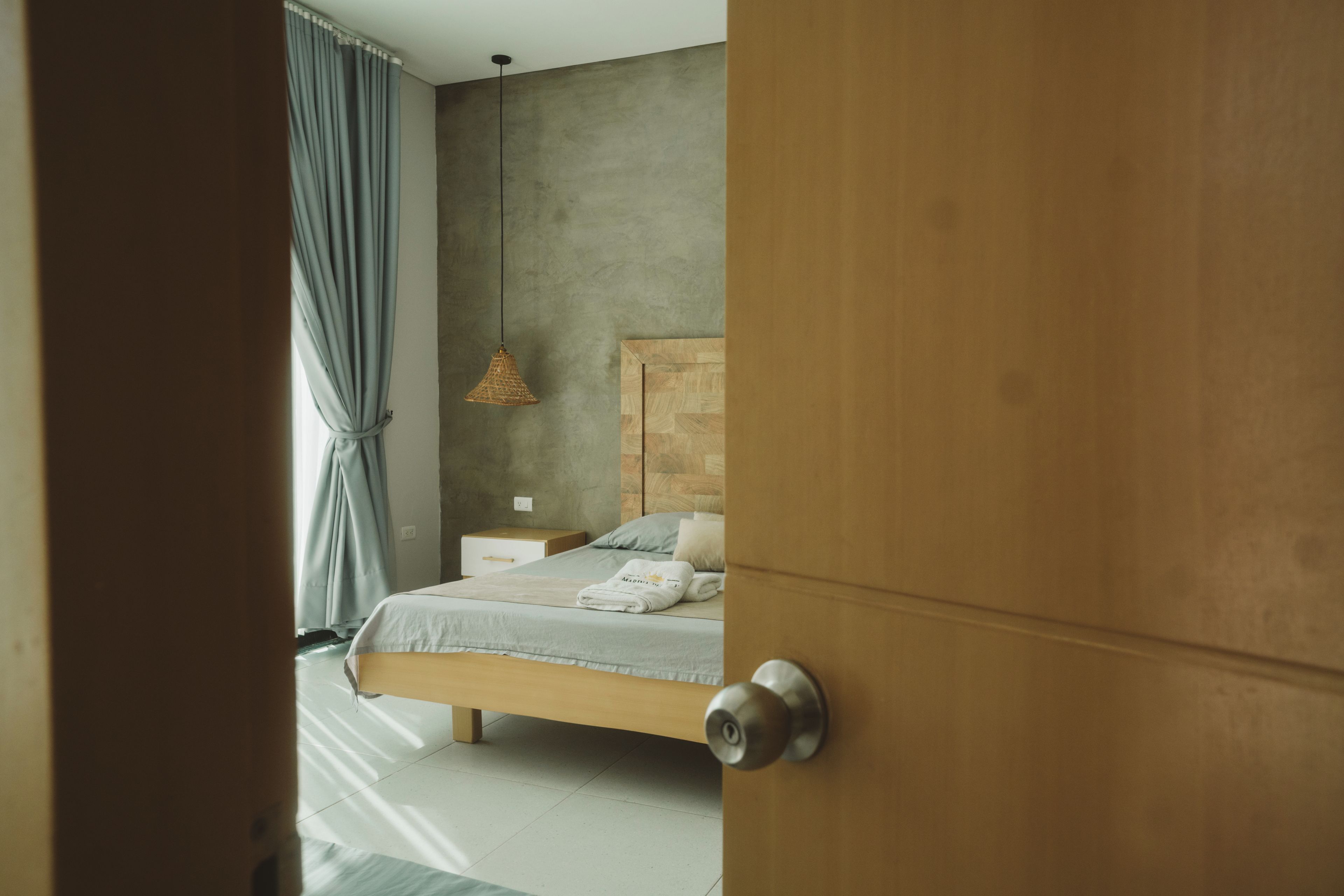 Un dormitorio con puerta de madera, una cama con cabecera, una mesita de noche, una lámpara colgante y una ventana con cortinas.