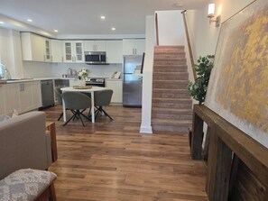 A modern kitchen with white cabinets, a microwave, and a refrigerator. There is a dining area with a table and chairs. A staircase leads to another level. - Furnished 3BR Home | All-Inclusive Monthly Vanier (Ottawa)