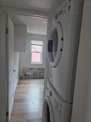 A laundry room with stacked washing machines, a radiator, and a window with blinds. - Furnished 3BR Home | All-Inclusive Monthly Vanier (Ottawa)