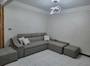 A modern living room with a grey sectional sofa, two matching ottomans, and a unique ceiling light fixture. - Family Apartment Near 45 Bridge, 200m from the Beach (Alexandria Governorate)