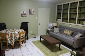 Una habitación con una mesa de comedor, sillas, un sofá, una mesita, una lámpara de pie, un radiador y una estantería.
