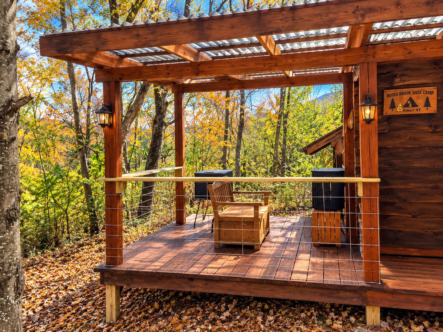Une terrasse en bois avec un banc et un barbecue, entourée d’arbres aux feuilles d’automne, et un panneau indiquant « Base camp de Roses Brook ».