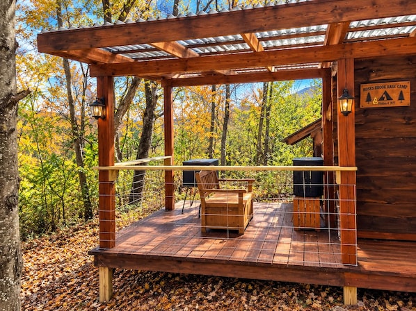 A wooden deck with a bench and a grill, surrounded by trees with autumn leaves, and a sign indicating 'Roses Brook Base Camp.'
