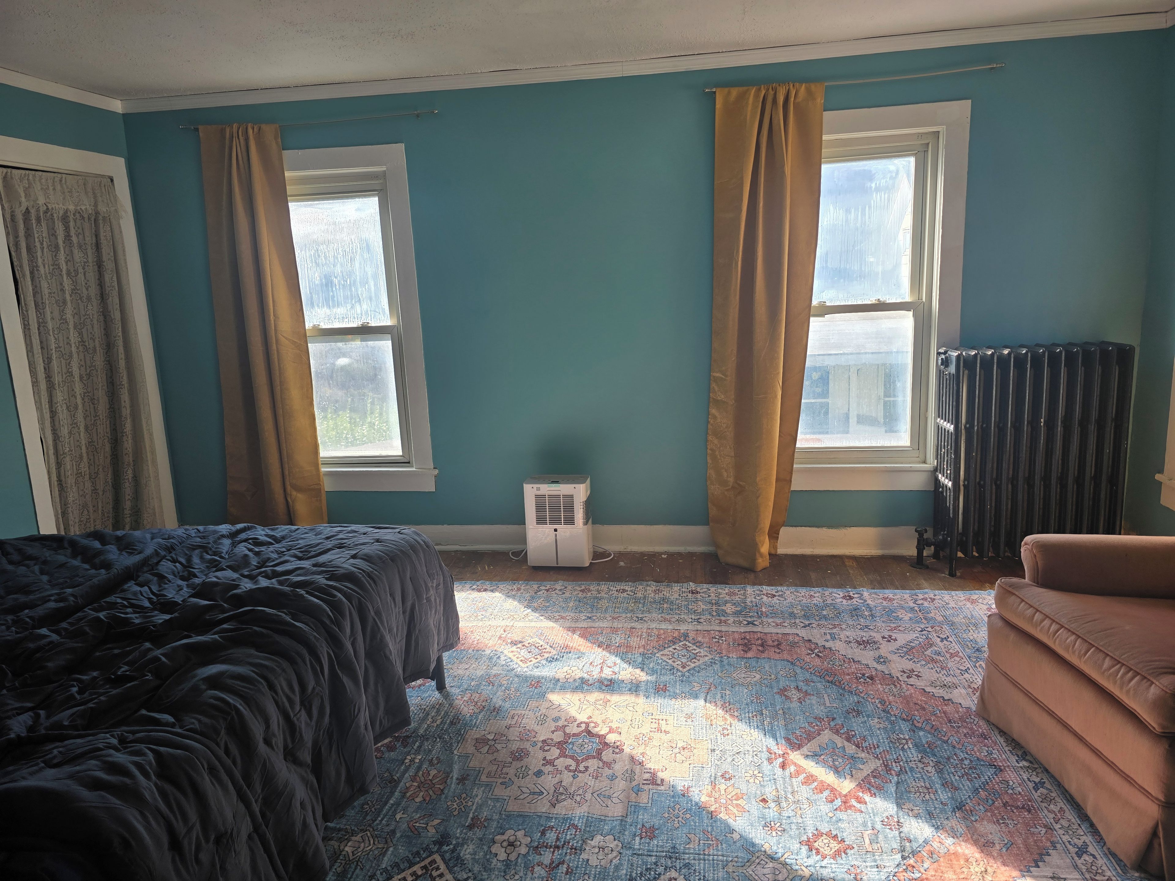 A room with a bed, a sofa, a radiator, and two windows with curtains.