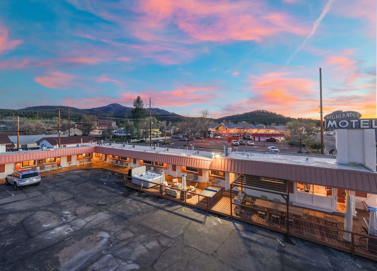 A motel with a covered outdoor seating area, a parking lot, and a mountainous backdrop.