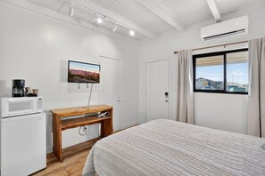 Een slaapkamer met een bed, een houten bureau, een witte koelkast, een televisie aan de muur en een raam met gordijnen.