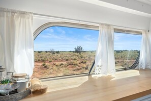 A room with a large window, a wooden table, and a view of a desert landscape. - Glamping Bus | Milky Way Views (Williams)