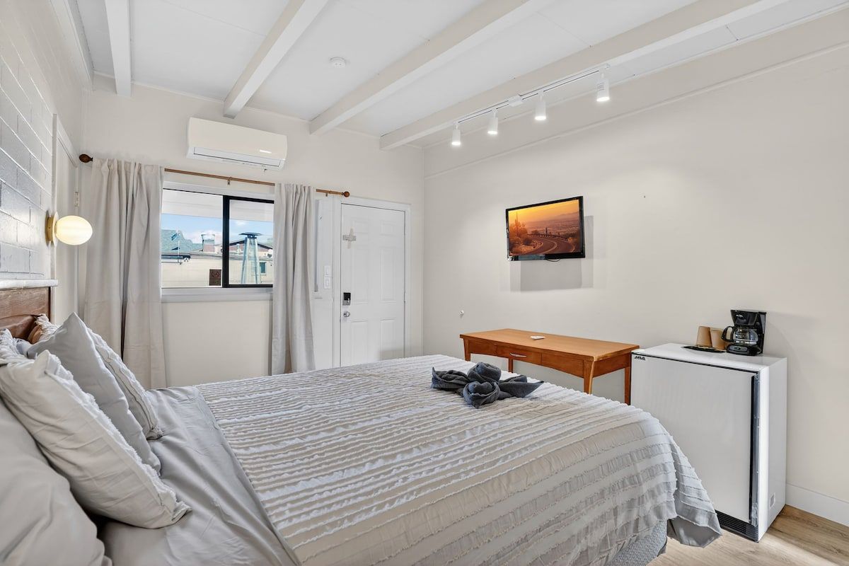 Un dormitorio con cama, escritorio, cafetera y un cuadro en la pared.