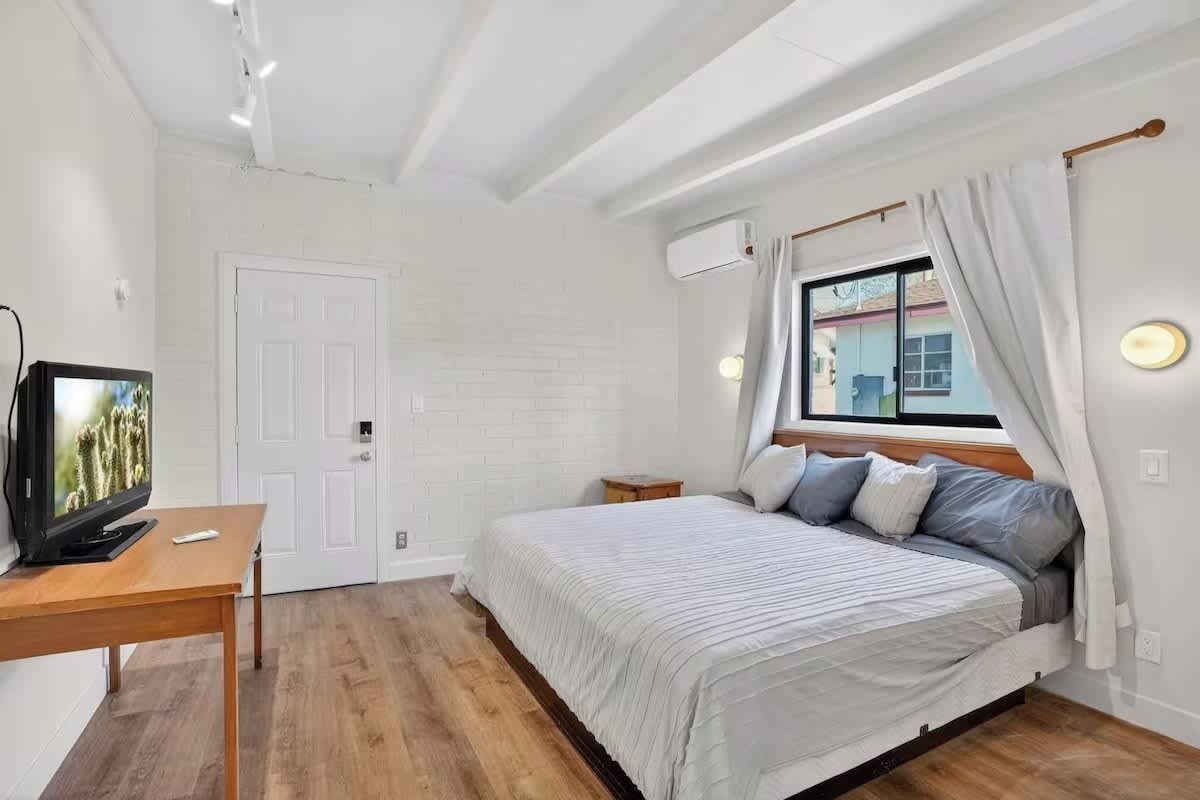 Ein Schlafzimmer mit einem Bett, einem Fernseher, einem Fenster mit Vorhängen und einer Klimaanlage.