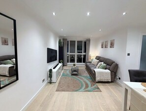 A modern living room with a flat-screen TV, a grey sofa, a coffee table, and a large mirror on the wall. - Tower Bridge Stay | 2 Bed | Fast WiFi & Parking (Greater London)