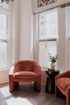Ein Zimmer mit zwei großen Fenstern, einem einzelnen orangefarbenen Sessel, einem kleinen Tisch mit einer Pflanze und einem weiteren Sessel.