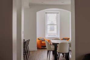 A dining area with a round table and chairs, a sofa with cushions, and a window with blinds. - Designer Combo Loft 4E & 4W|Gold Coast/River North (Chicago)