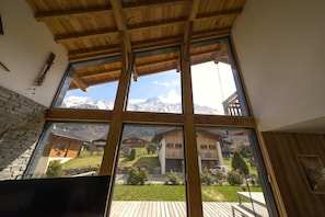 Una habitación con un ventanal grande que da a un paisaje montañoso, vigas de madera en el techo y una pared de piedra.