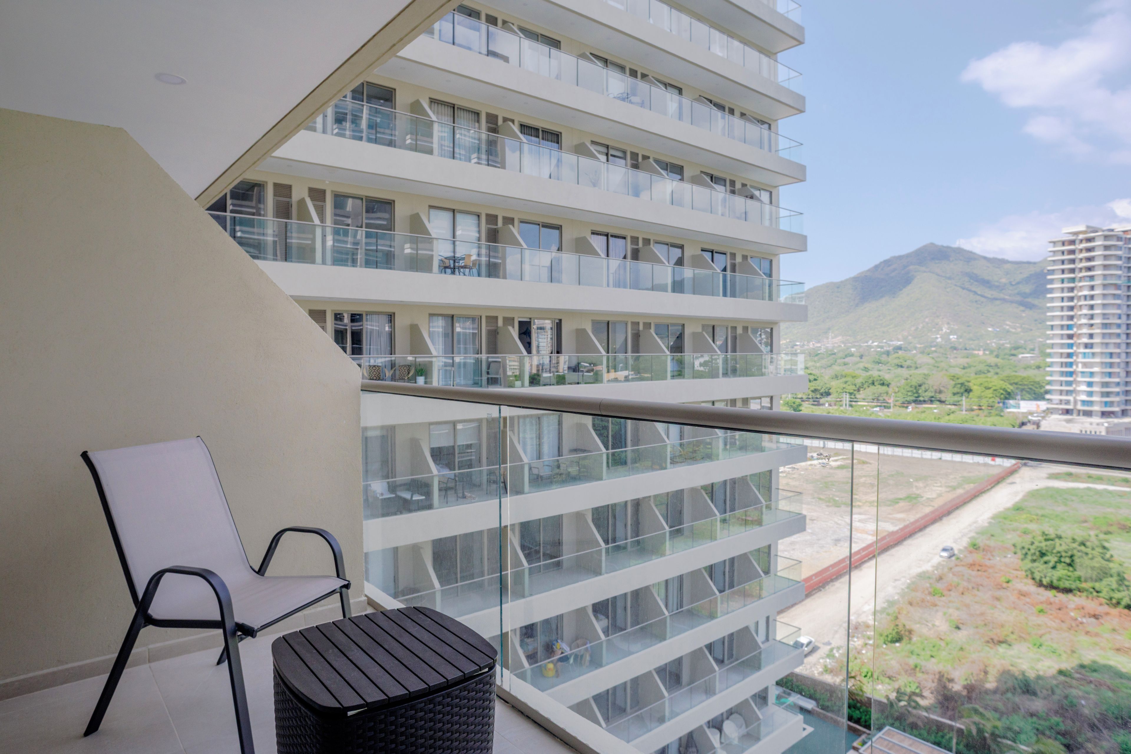 Un balcon avec une chaise et une petite table, donnant sur un immeuble moderne doté de nombreux balcons, avec un paysage montagneux en arrière-plan.