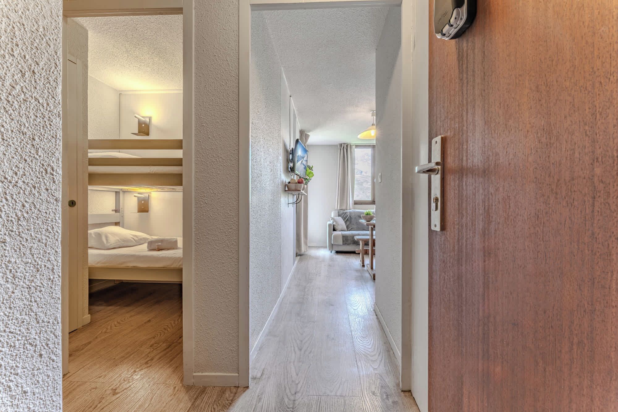Un pasillo de hotel con suelo de madera, una habitación con literas a la izquierda, un área de estar con una mesa y sillas, y una puerta de madera a la derecha.
